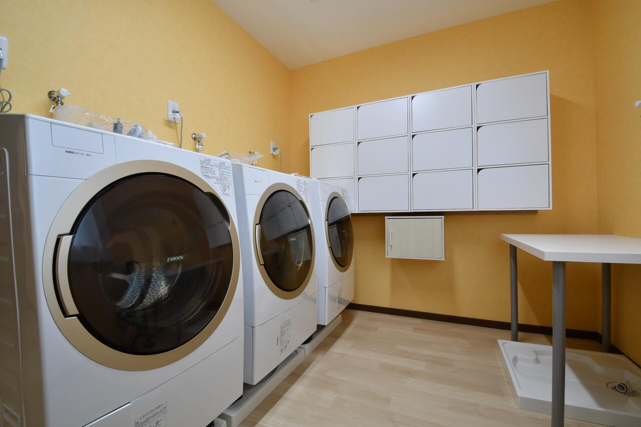 ランドリースペースの様子。ドラム式洗濯機が3台並んでいます。洗濯機の脇の壁に専有部ごとの収納が設置されています。|2F ランドリー